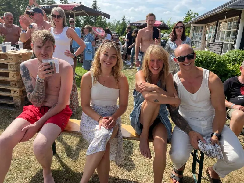 Fire smilende gæster sidder sammen på en bænk i solen på festivalpladsen.